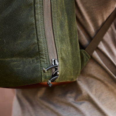 Close-up of an individual sporting a GORUCK Bullet Ruck - Waxed Canvas backpack, showcasing the textured green material and visible zipper. The backpack is worn over a grey shirt, with the image focusing on its detailed design reminiscent of a retro canvas rucksack and highlighting part of the shoulder strap.