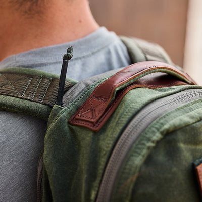 A person in a gray shirt carries the GORUCK Bullet Ruck - Waxed Canvas, highlighting its green waxed canvas complemented by brown leather accents. The emphasis is on the shoulder strap and top handle, showcasing the retro design and meticulous stitching details of this rucksack.