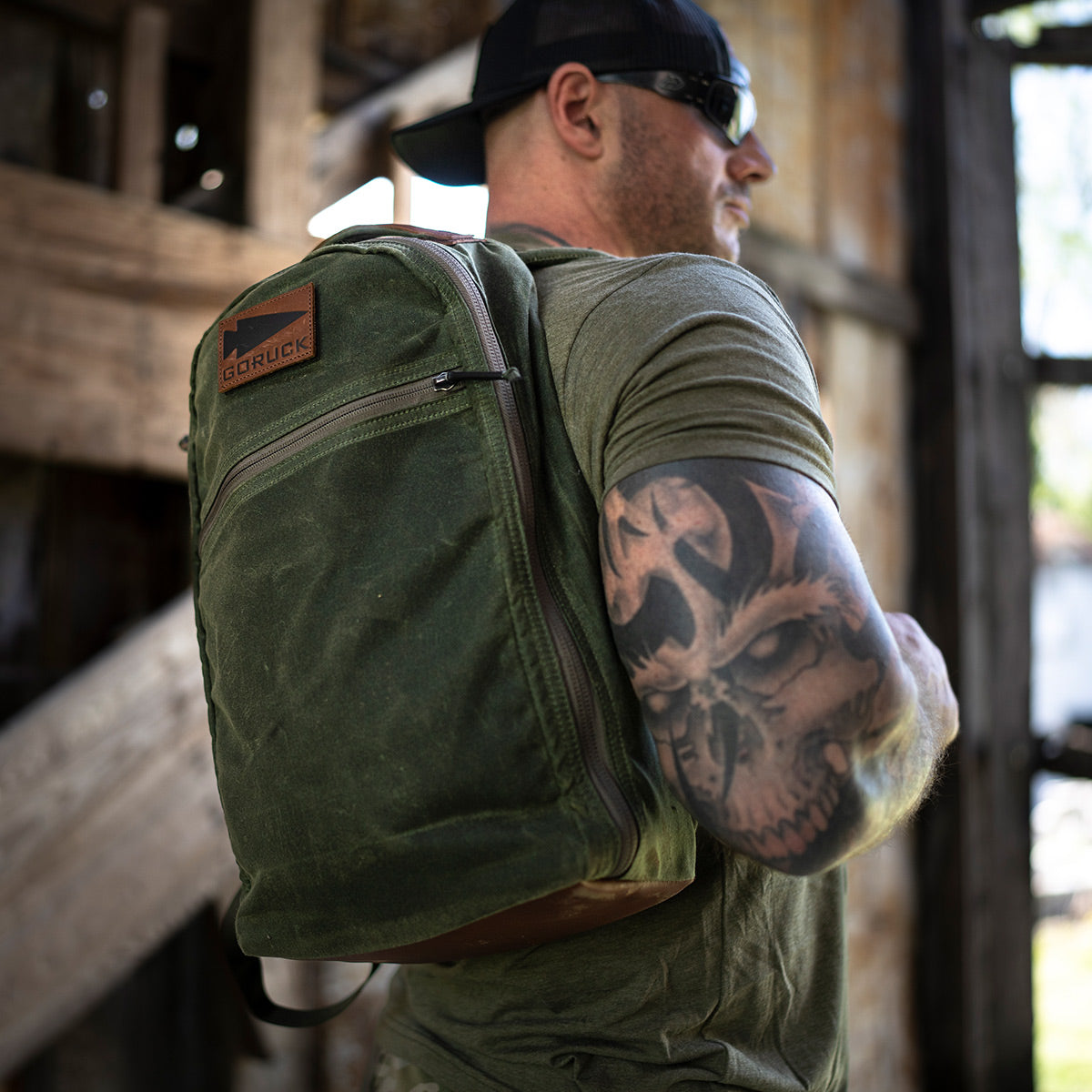 A man wearing sunglasses, a cap, and a green shirt stands in a rustic building. He carries a Bullet Ruck - Waxed Canvas from GORUCK on his shoulder. His arm is tattooed with an intricate skull design as sunlight filters through the wooden structure.