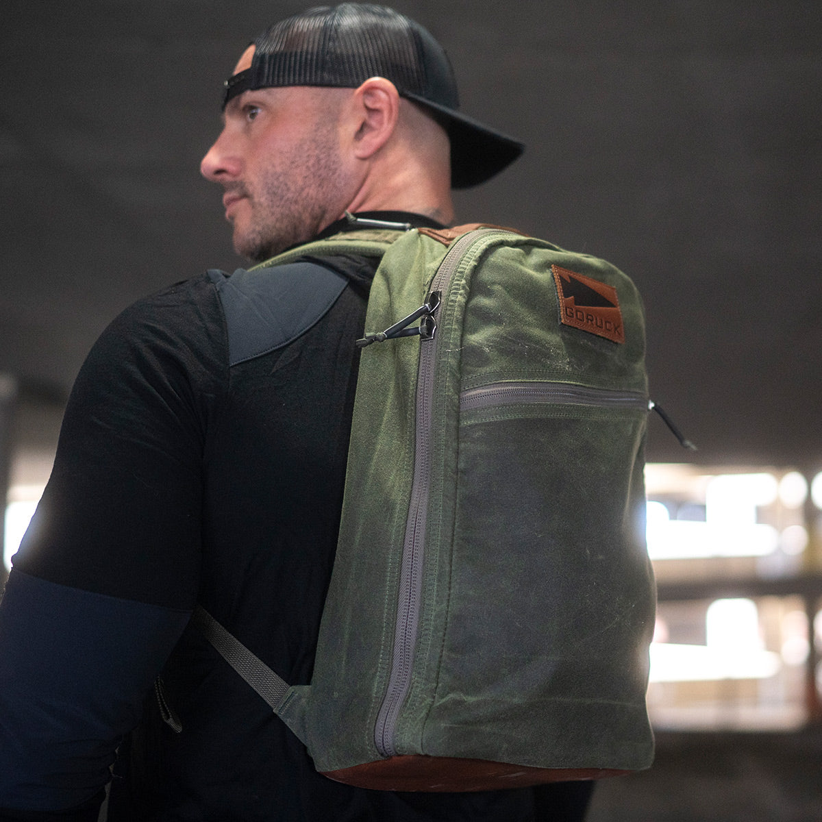 A man wearing a black cap and black shirt carries a green Bullet Ruck - Waxed Canvas from GORUCK over one shoulder. The partially visible logo on the rucksack reads GORUCK. The blurred background suggests an indoor setting.