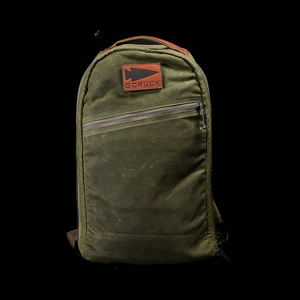 A waxed canvas GORUCK Bullet Ruck backpack in green, featuring a brown leather logo patch on the upper front and a front zipper pocket, isolated against a black background.