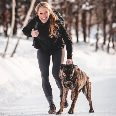 A woman joyfully runs with her dog on a snowy path surrounded by trees, her loyal companion keeping pace beside her Bullet Ruck Classic - Cordura, ideal for any rucking adventure.