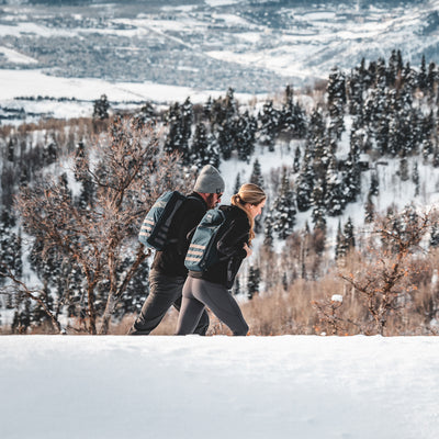 Two people trek uphill through the snowy landscape with snow-covered trees and mountains behind them, each carrying a durable Bullet Ruck Classic - Cordura daypack.