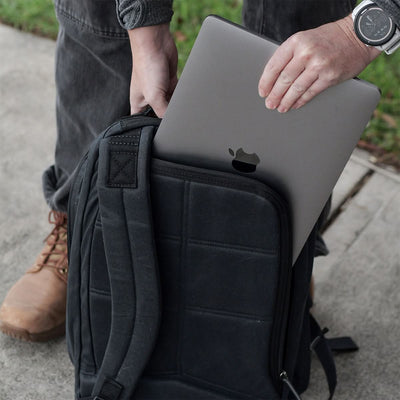 A person places a laptop into a black Bullet Ruck - Waxed Canvas by GORUCK. The scene unfolds outdoors on a sidewalk, as the individual, donned in a gray jacket, a watch, and brown boots, seamlessly blends practicality with timeless style.