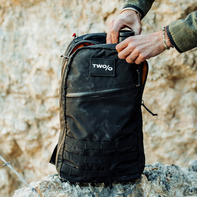 Amidst rugged rocky terrain, someone opens a Bullet Ruck - Ripstop ROBIC®, Michael Easter edition, featuring a "TWO/O" label.