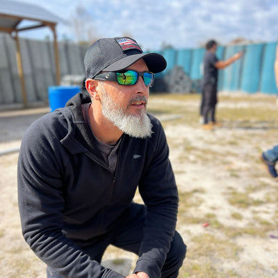 A man in sunglasses and a Cadre Hat - CORDURA® kneels outdoors, surrounded by blurred figures and a fence, symbolizing the resilience of a Special Forces Cadre.