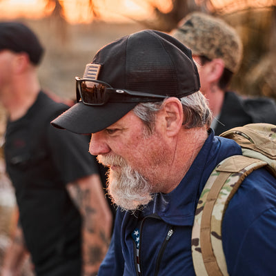 A bearded man wearing a Cadre Hat - CORDURA® and sunglasses, with a durable 500D CORDURA® backpack, walks outdoors alongside two others.