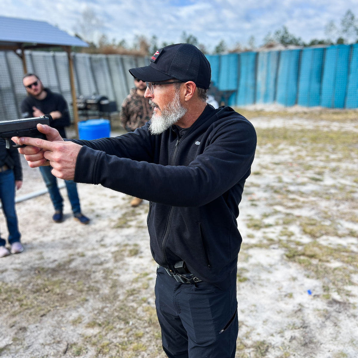 A man in a Cadre Hat - CORDURA® targets his handgun at an outdoor shooting range, with onlookers watching intently.