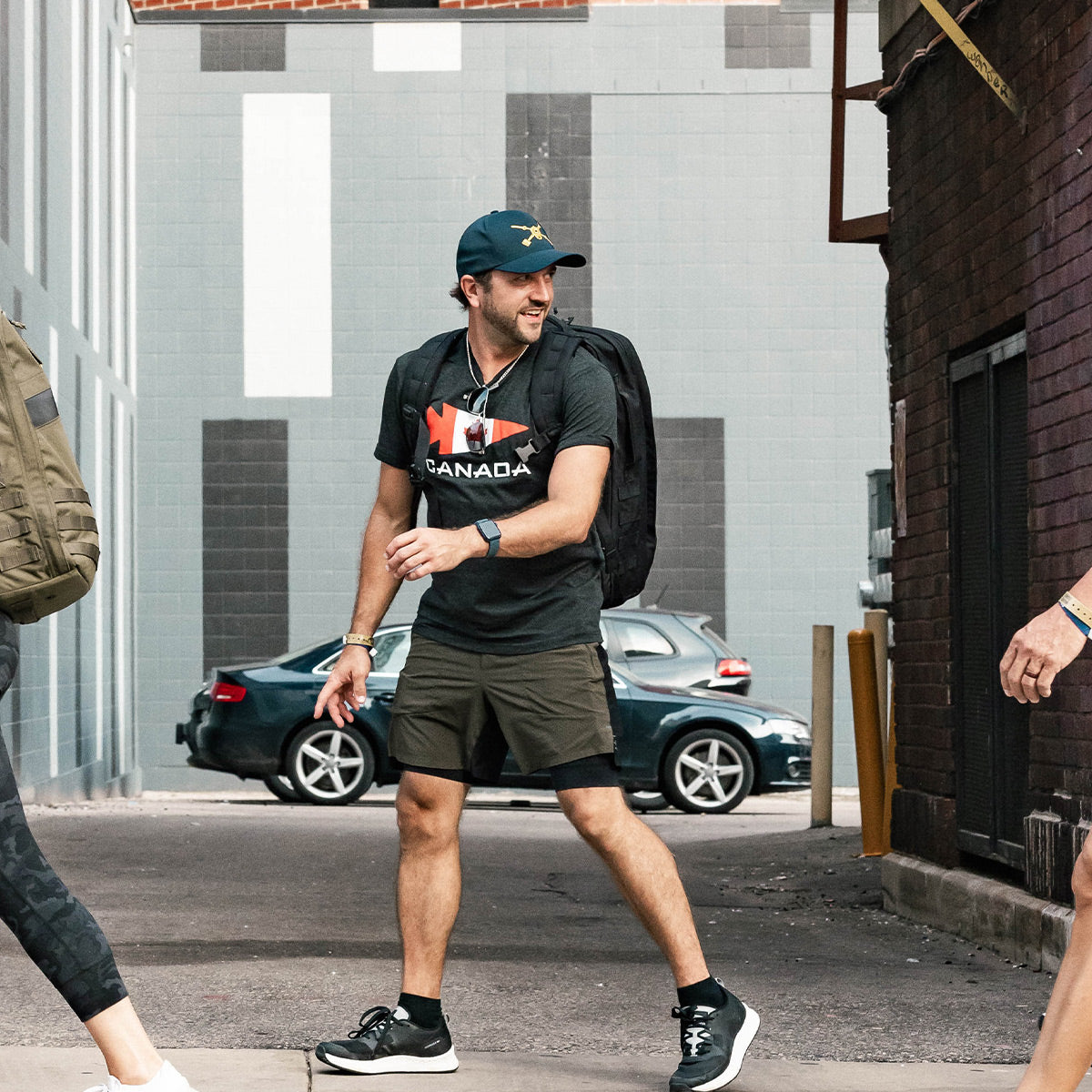 A man in a GORUCK Canada Tee - Tri-Blend and black cap walks outdoors, carrying a GORUCK backpack. He's in motion, navigating through the cityscape of brick and painted walls. A sleek black car rests in the background, matching his adventurous vibe.