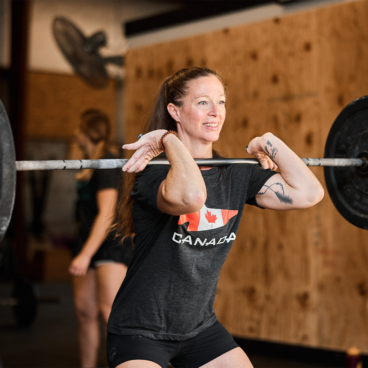 A woman with a ponytail is performing a front squat at the gym, lifting a barbell in her GORUCK Canada Tee - Tri-Blend from vendor-unknown. Her black T-shirt displays the Canadian flag design and is made from high-quality tri-blend material. In the background, another person is exercising.