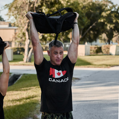 A man with short gray hair lifts a GORUCK backpack above his head while outdoors. He wears a GORUCK Canada Tee - Tri-Blend, which showcases a Canadian flag along with the word CANADA and is made from high-quality tri-blend fabric. Tall trees and buildings can be seen in the background.