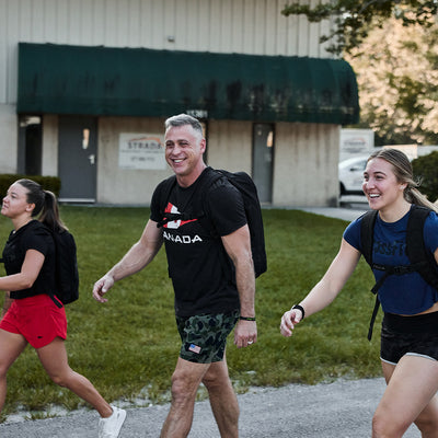 Three individuals clad in athletic gear and backpacks stride down the street, with the person in the middle showcasing a GORUCK Canada Tee crafted from premium tri-blend fabric. They're all smiles, embodying the essence of adventure. As they move forward as if on a GORUCK challenge, trees sway gently beside a building in the background.