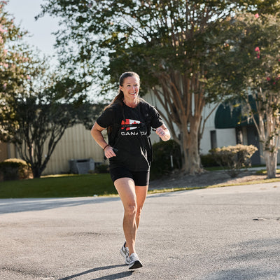 A woman is jogging on a sunny day, wearing a GORUCK Canada Tee - Tri-Blend from vendor-unknown. She smiles as she runs on a paved surface, with trees and a building in the background.