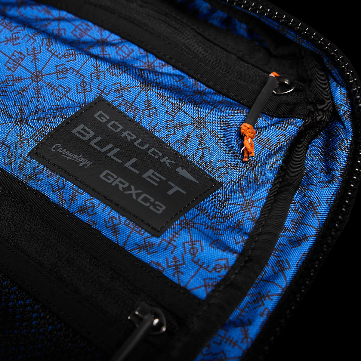 A close-up of a GORUCK backpack's interior shows blue fabric featuring a geometric pattern. A black patch displays "GORUCK Bullet Ruck x Carryology - Berserker Viking" with "GRXC3, Constellation." An orange zipper pull on the right pays homage to the Viking Bullet Ruck aesthetic, capturing the ideal fusion of durability and style from the GRXC program.