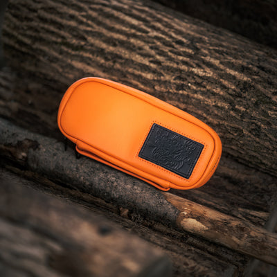 A GORUCK Echo Field Pocket x Carryology - Berserker Viking, featuring a vibrant orange color and a black embossed patch, sits atop a stack of rough-textured logs. Its design evokes the image of a Viking shield amidst the untamed landscape, with its texture and color creating a striking contrast against the natural wooden background.