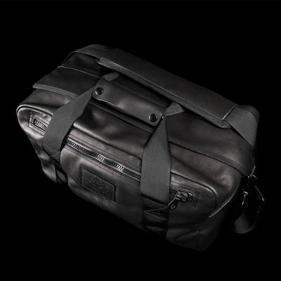 Introducing the Kit Bag x Carryology - Berserker Viking by GORUCK: a sleek black leather duffel bag inspired by a Viking kit bag, featuring sturdy handles and a detachable shoulder strap. The design includes a front zippered pocket and a logo patch, set against a black background.