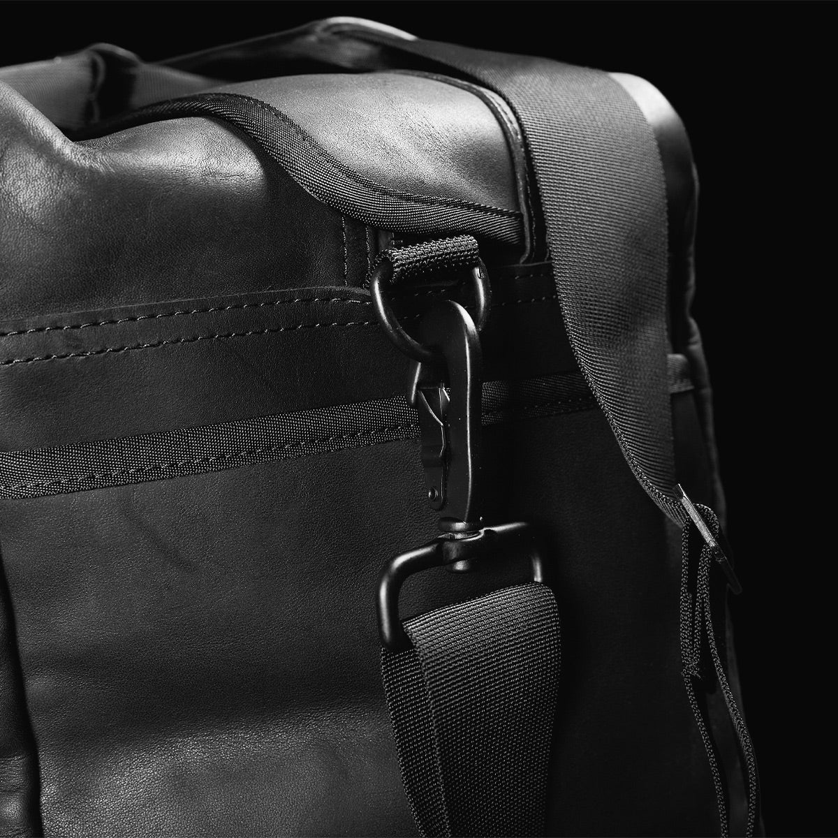 Close-up of the GORUCK Kit Bag x Carryology - Berserker Viking, featuring a sleek black leather finish with a textured surface reminiscent of Viking armor. The focus is on the sturdy metal clasp attached to a wide fabric strap, highlighting the bag's design and craftsmanship against a dark background, echoing the rugged style of Norsemen warriors.