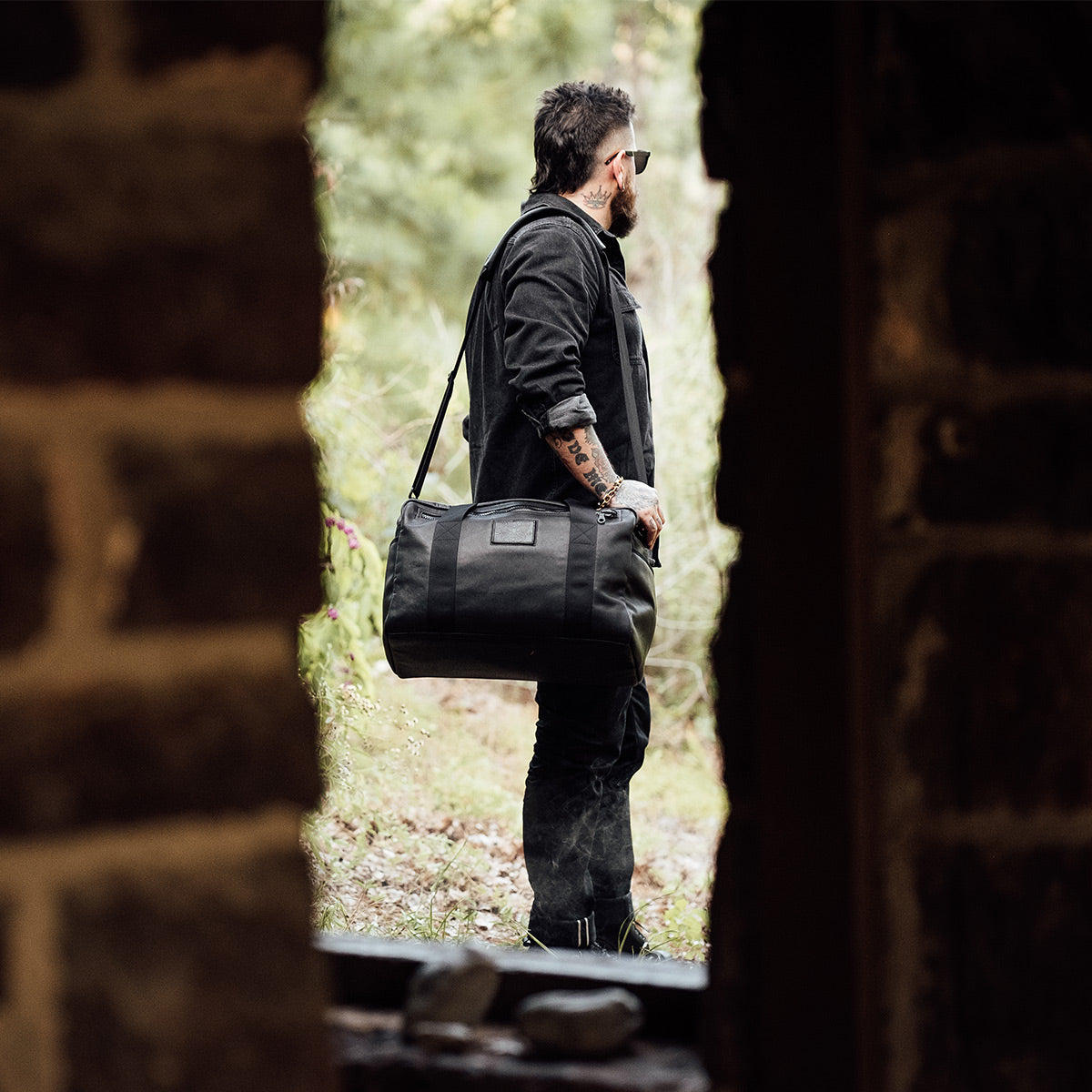 A person with tattoos and sunglasses stands outdoors, holding the GORUCK Kit Bag x Carryology - Berserker Viking. They are viewed through a rough stone doorway, with green foliage visible in the background, evoking a rugged, adventurous feel reminiscent of Norsemen warriors on their travels.