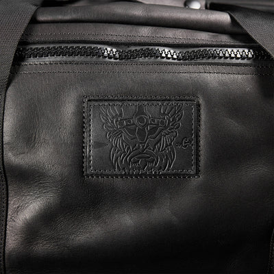 Close-up of the GORUCK Kit Bag x Carryology - Berserker Viking, a black leather bag reminiscent of a Viking kit bag. It showcases a textured, embossed patch with an intricate design of a stylized face wearing a winged helmet, along with a visible zipper above the patch that embodies the spirit of Norsemen warriors.