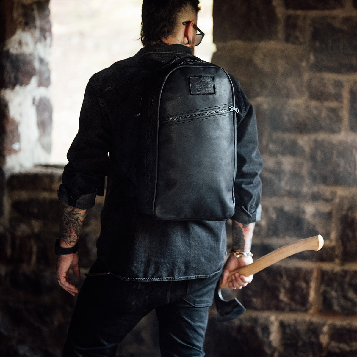 A person dressed in dark attire stands with their back to the camera, holding an axe and wearing the GORUCK's Bullet Ruck x Carryology - Berserker Viking backpack. They are in a rustic environment with stone walls, and tattoos visible on their arms.