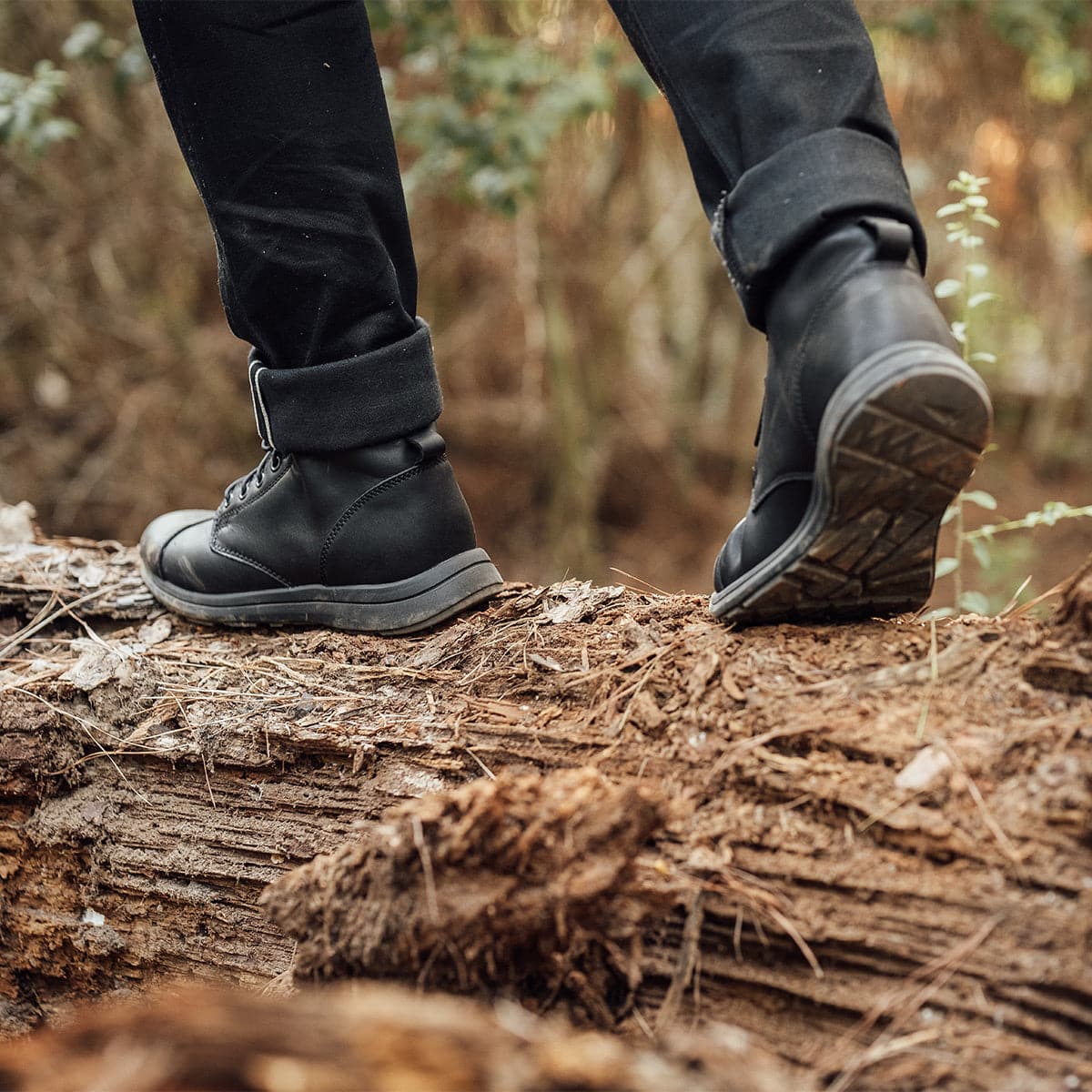 A person wearing the GORUCK MACV-1 Traveler x Carryology - Berserker Viking boots and black pants strides confidently across a fallen tree trunk in a forest, with a backdrop of blurred greenery.