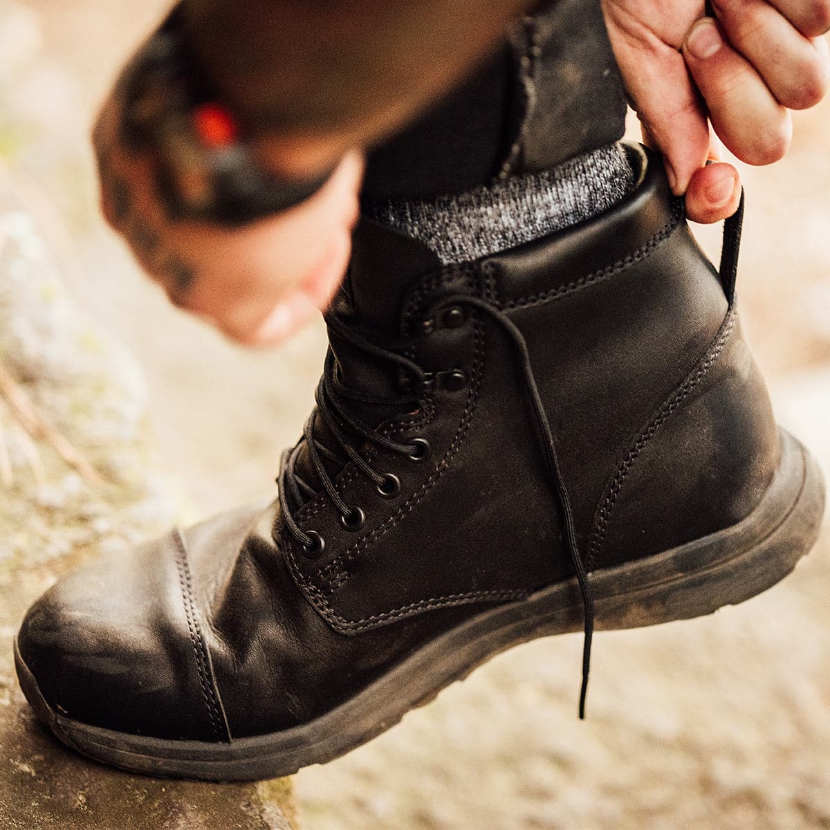 A person wearing the GORUCK MACV-1 Traveler x Carryology - Berserker Viking boots is adjusting the laces. The focus is on the boot, with hands pulling the laces tight. Designed for travel and adventure, these water-resistant leather boots are set against an outdoor natural setting.