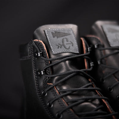 Close-up of the black GORUCK MACV-1 Traveler x Carryology - Berserker Viking boots, featuring intricate stitching and laces against a dark backdrop. The emphasis is on the texture of the water-resistant leather and design, spotlighting a logo patch with an arrow and abstract pattern on the tongue, perfect for travel and adventure.