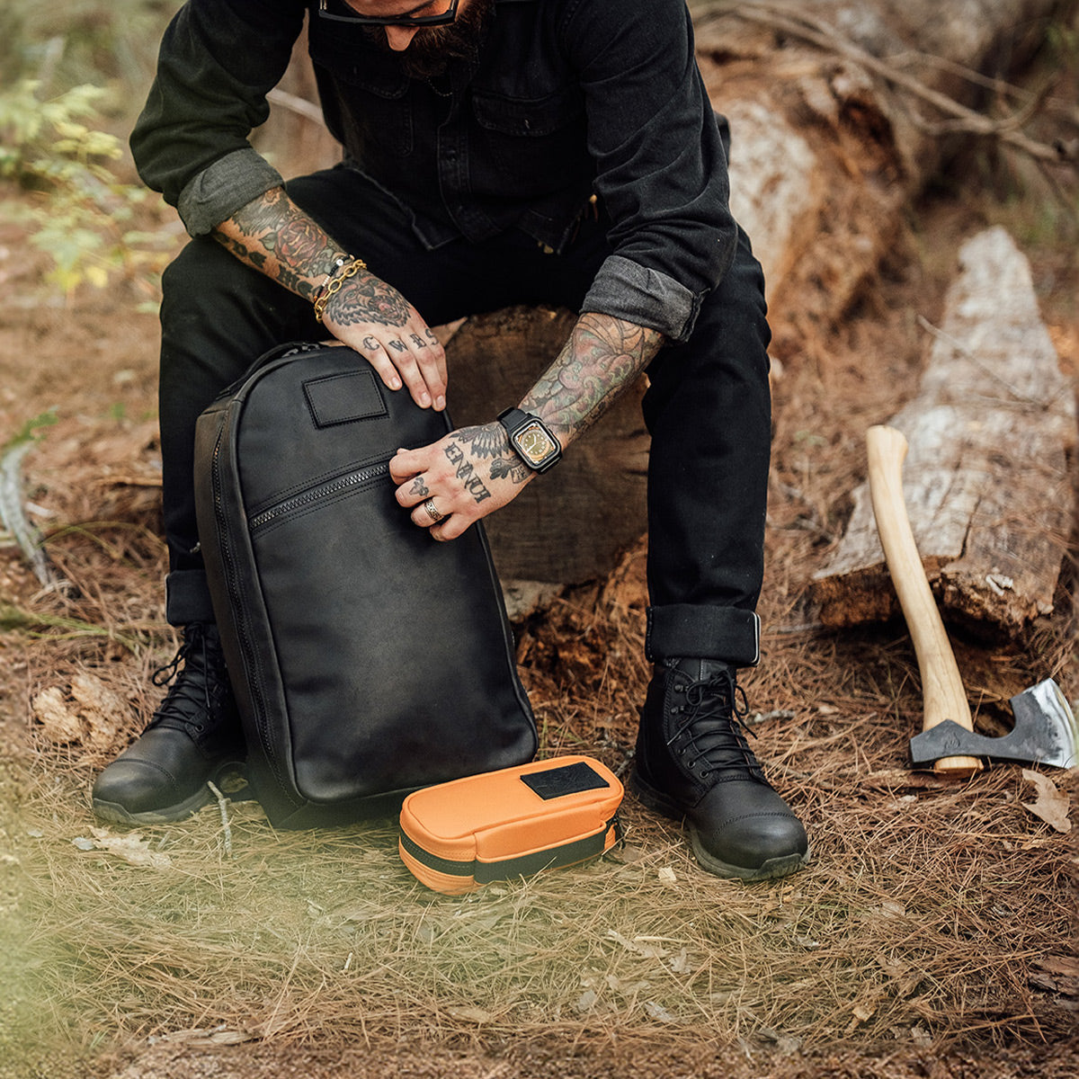 A tattooed individual lounges on a log in the forest, embodying a contemporary Norseman style with black boots and attire. Beside them, a GORUCK Echo Field Pocket x Carryology - Berserker Viking backpack rests alongside an orange case on the ground, while an axe that evokes the weaponry of Vikings leans against another log.