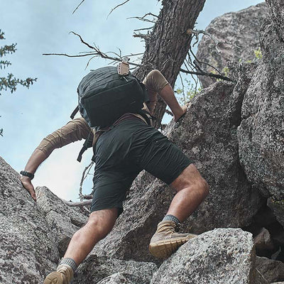 A person clad in GORUCK’s Men’s Challenge Shorts crafted from lightweight ToughDry® fabric and wearing a backpack is rock climbing on rugged terrain, reaching towards the sky. The scene unfolds in a natural setting with a cloudy sky as the backdrop.