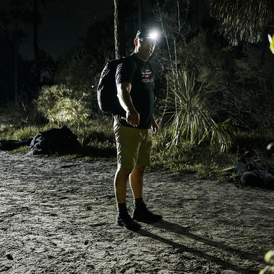 In a dimly lit outdoor setting, a man is highlighted by the glow of a headlamp. He's clad in a black shirt and khaki GORUCK Men’s Challenge Shorts - Lightweight ToughDry®, complete with cargo pockets, while carrying a backpack. The scene is enhanced by the presence of trees and bushes, contributing to the nighttime ambiance.