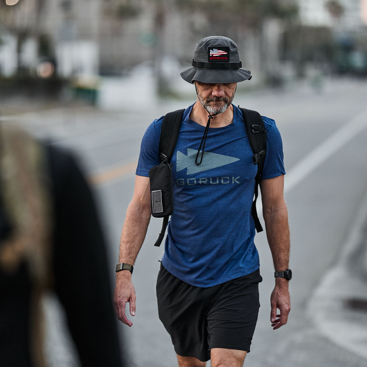 A man strolls outdoors in a blue shirt and black shorts, sporting a Boonie Hat - Tactical - ToughDry and carrying a backpack with a bottle attached.