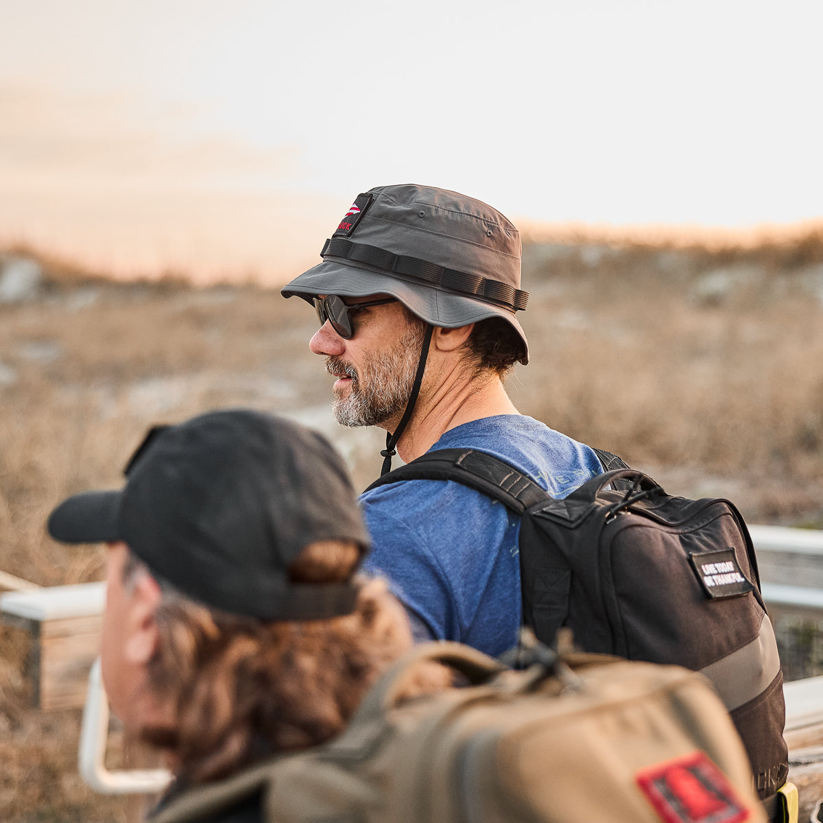 Two people in Tactical ToughDry boonie hats and backpacks are outdoors. One gazes ahead, evoking a military style, with gear ready for any adventure against the backdrop of grass and open sky.