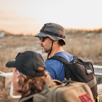 Two people in Tactical ToughDry boonie hats and backpacks are outdoors. One gazes ahead, evoking a military style, with gear ready for any adventure against the backdrop of grass and open sky.