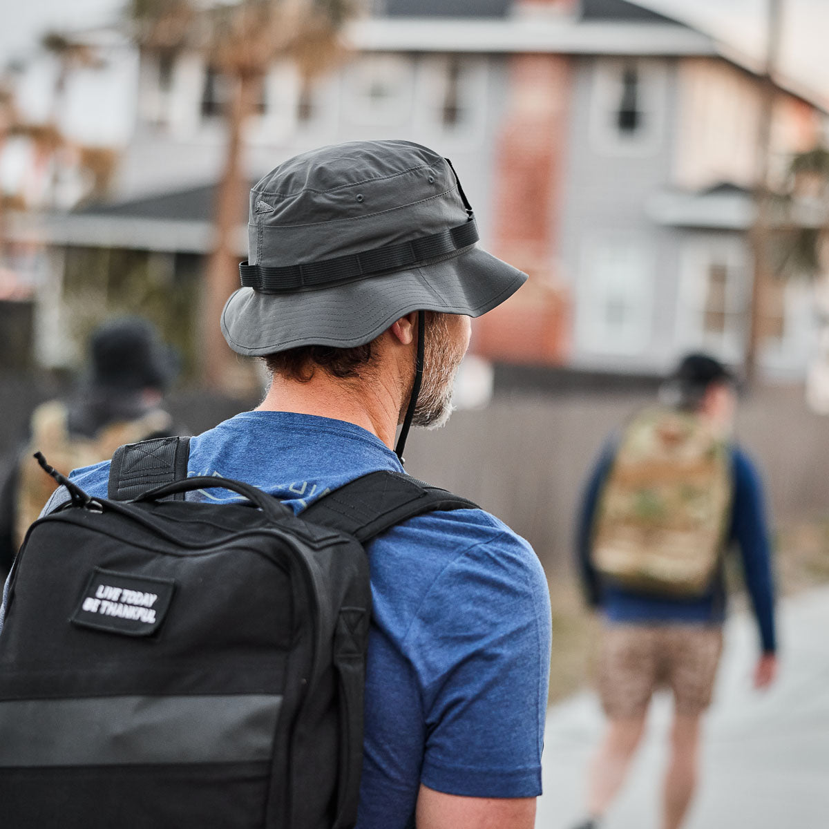 A man in a blue shirt and ToughDry Tactical Boonie Hat, with a backpack, strides down the street, blurred houses in the background.