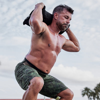 A man with a bare upper body is working out outside, doing squats while balancing a weight bag on his shoulders. He's sporting GORUCK's Performance Briefs, designed for moisture-wicking performance and style against the backdrop of a partly cloudy sky, ensuring he stays comfortable as he powers through his workout.