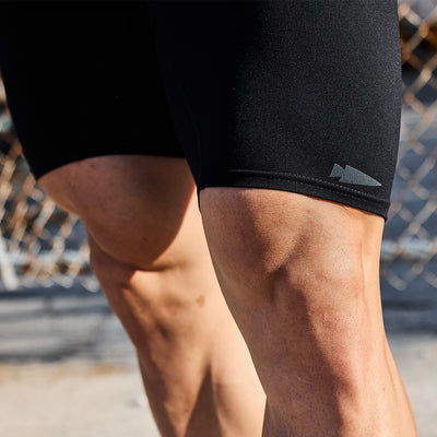 Close-up of a person's muscular legs in GORUCK's Performance Briefs. A small arrowhead symbol is visible on the breathable fabric. In the background, a chain-link fence appears blurred.