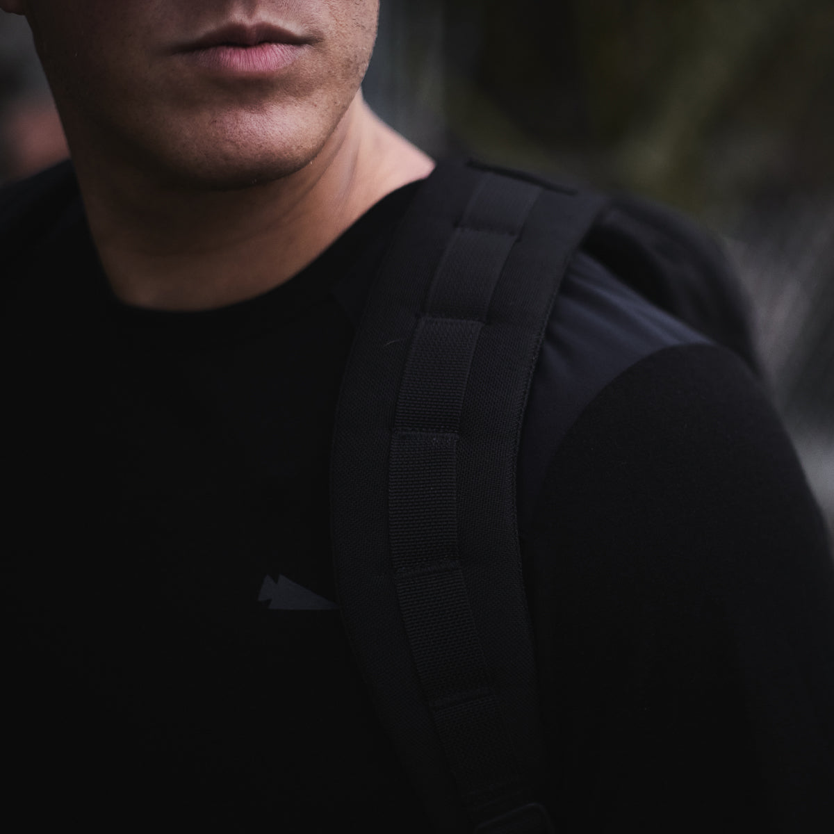 A person in a black shirt and backpack emerges from a dark, blurred backdrop, their outfit subtly echoing the rugged charm and blend of function with style found in the Men’s Commando Long Sleeve - Merino Wool.