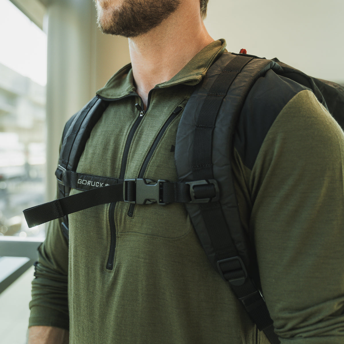 Indoors, a person in a green shirt similar to the Men’s Commando Half Zip - Merino Wool wears a backpack with reinforced paneling and a strong sternum strap.