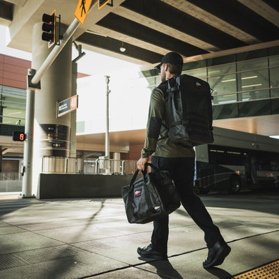 Wearing a Men's Commando Half Zip - Merino Wool, and carrying both a backpack and duffel bag, an individual navigates an urban transit area, weaving between buses under the bridge.