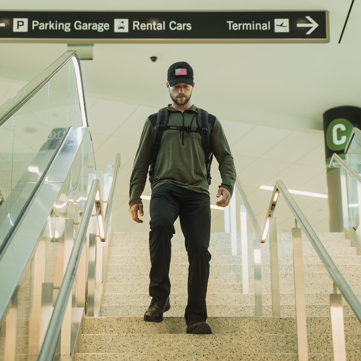 A man in a Men’s Commando Half Zip - Merino Wool sweater, featuring reinforced paneling, descends airport stairs beneath signs for parking, rental cars, and the terminal.