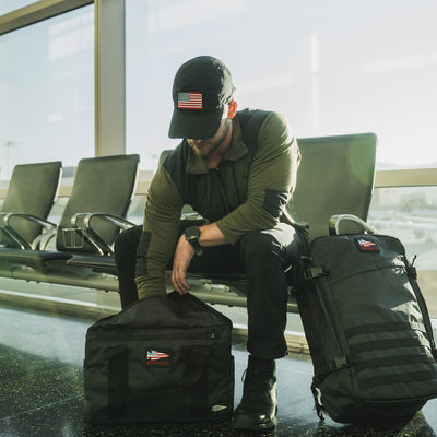 A man in a cap with a U.S. flag sits at the airport, organizing his black bags. Dressed in the Men’s Commando Half Zip - Merino Wool sweater, he enjoys the sunlight streaming through large windows.