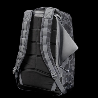 The GR1 USA - Cordura (The Original Ruck), a gray camouflage backpack with padded straps, resembles a Special Forces rucksack, featuring a laptop partially visible in the side zipper pocket against a black background.