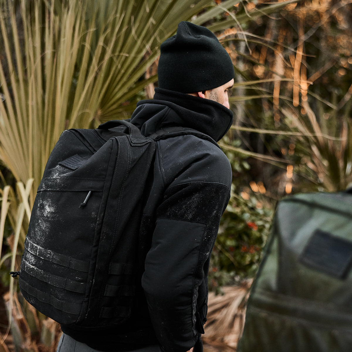 A person in a Men's Task Force Dagger Jacket - Polartec Fleece, wearing a black beanie and carrying a black backpack, stands outdoors near lush foliage, evoking Special Forces resilience.