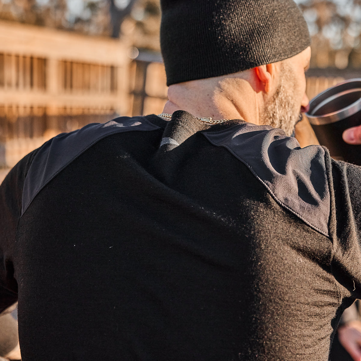 A man in a black beanie drinks from a mug outdoors, wearing a Men’s Commando Long Sleeve - Merino Wool shirt, with a wooden fence behind him.