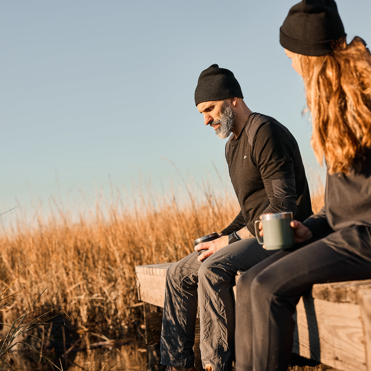 Two people in black clothing sit on a wooden dock, holding mugs, with tall grass and clear skies behind them. One is wrapped in a cozy Men’s Commando Long Sleeve made of Merino Wool, ideal for breezy days by the water.