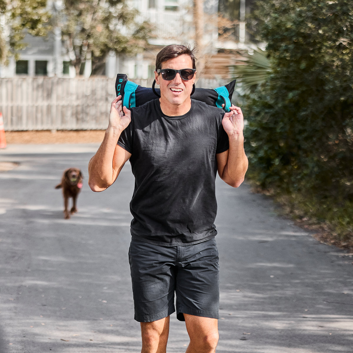 A man in sunglasses, carrying a sandbag on his shoulders, walks down the street wearing Men’s Simple Shorts made of lightweight ToughDry® fabric. A brown dog lingers in the background, seemingly impressed by the gear's resilience.