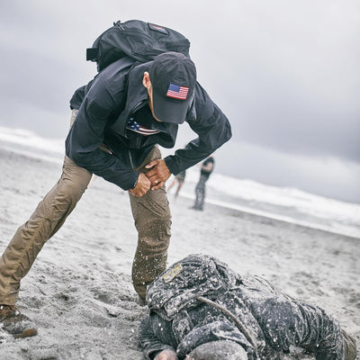 Under a cloudy sky, someone in tactical gear with roomy cargo pockets assists another on the sand. Their Men’s Challenge Pants made with Lightweight ToughDry® fabric promise durability in challenging beach conditions.