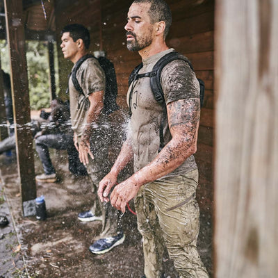 Two men with bulging backpacks stand under a wooden shelter being sprayed with water. Wearing Men’s Challenge Pants crafted from abrasion-resistant Lightweight ToughDry® fabric, they enjoy durable protection in the elements while their fast-drying pants shed rain and mud.