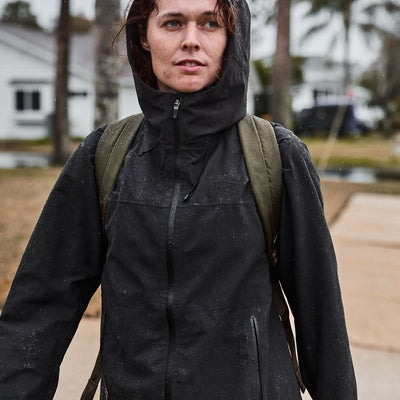 The individual is equipped with a hooded black rain jacket and a green Packable Bullet Ruck made of Ripstop ROBIC® nylon, ready to face the rainy day.