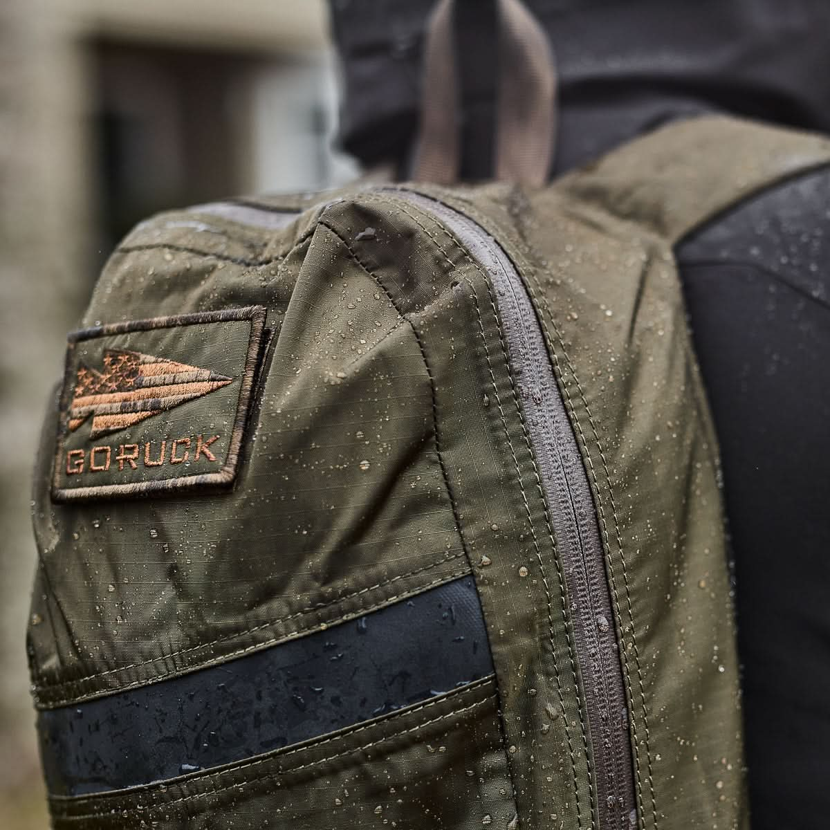 Close-up of an olive-green Packable Bullet Ruck - Ripstop ROBIC® backpack, showcasing raindrops and a patch logo, worn on the back of a person in a dark jacket.